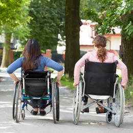 Two women wheeling their manual wheelchairs down a path in the park. 