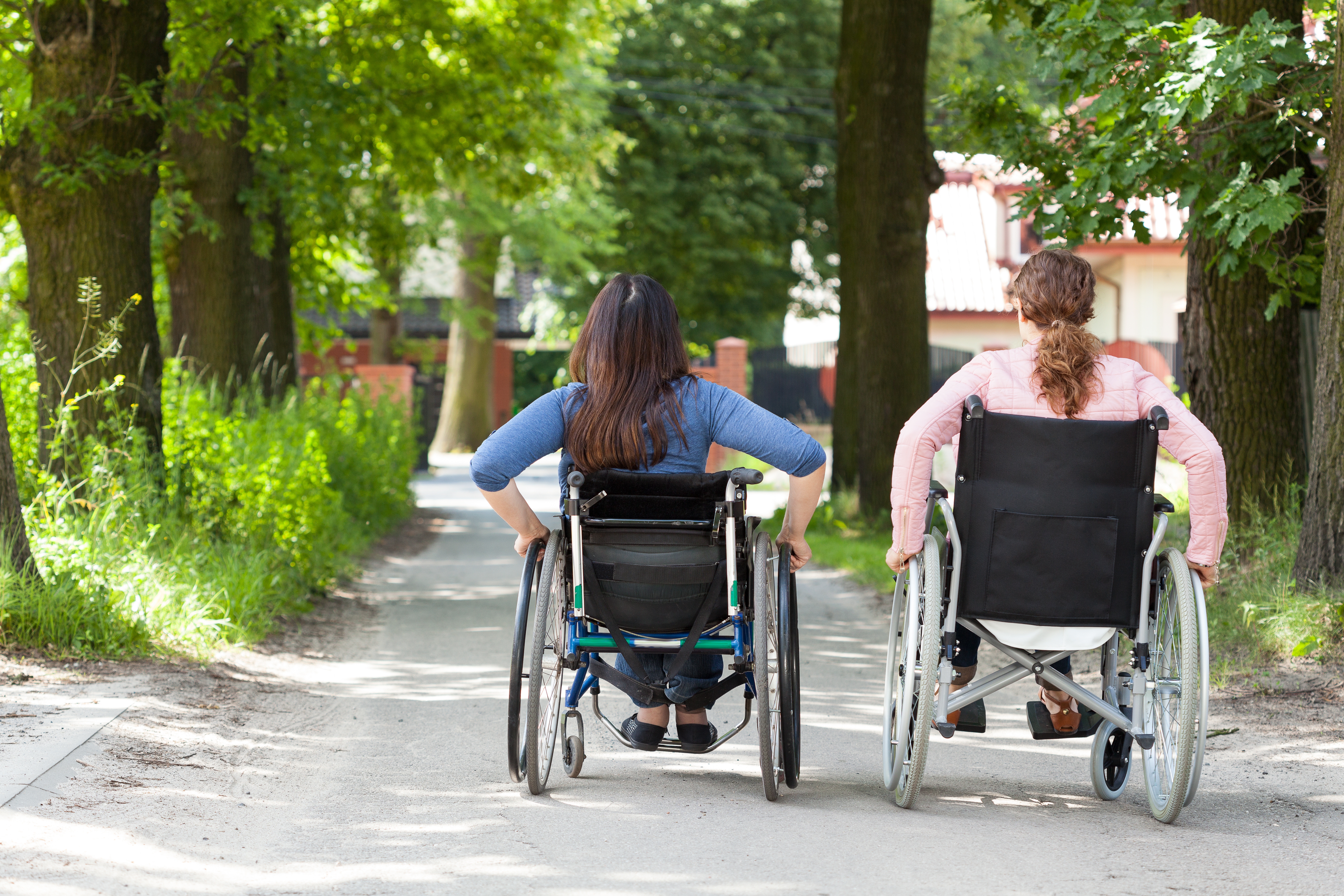 Two women wheeling their own manual wheelchairs down a dirt path through trees in a park.