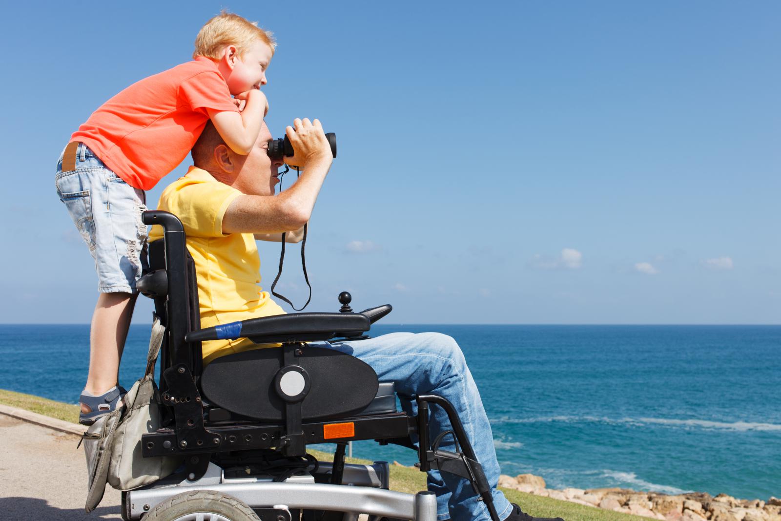 this is an image of man in his power wheelchair alongside ocean looking through binoculars with his son on his back
